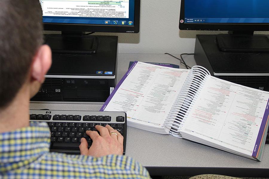 An individual sits at a computer, typing on the keyboard, 他们旁边的桌子上放着一本医学编码信息的书
